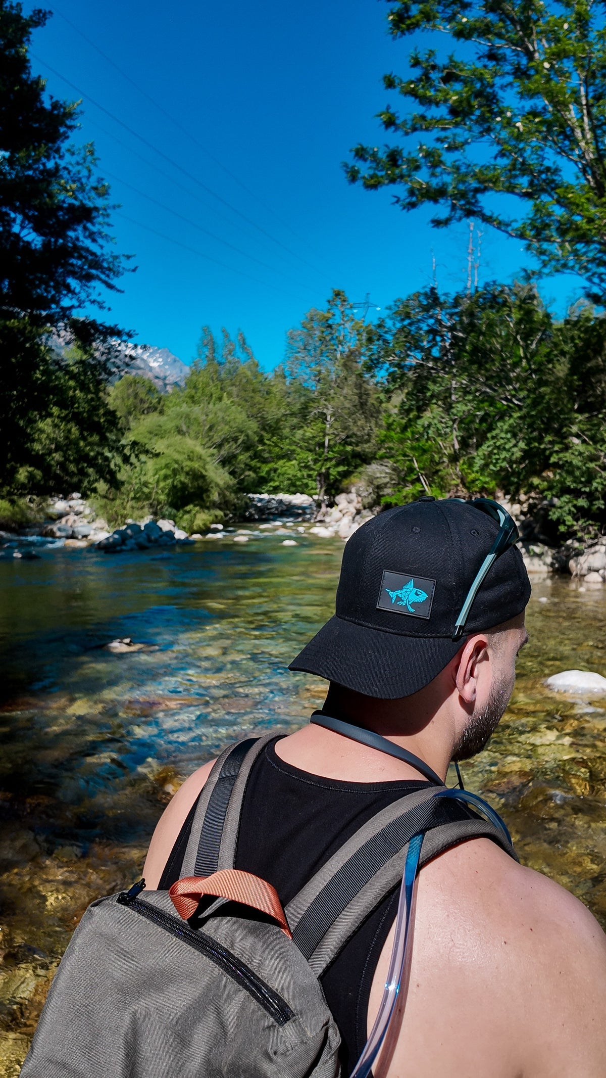 Connected Carper black baseball cap with electric blue predator fish patch worn by a man near a mountain river