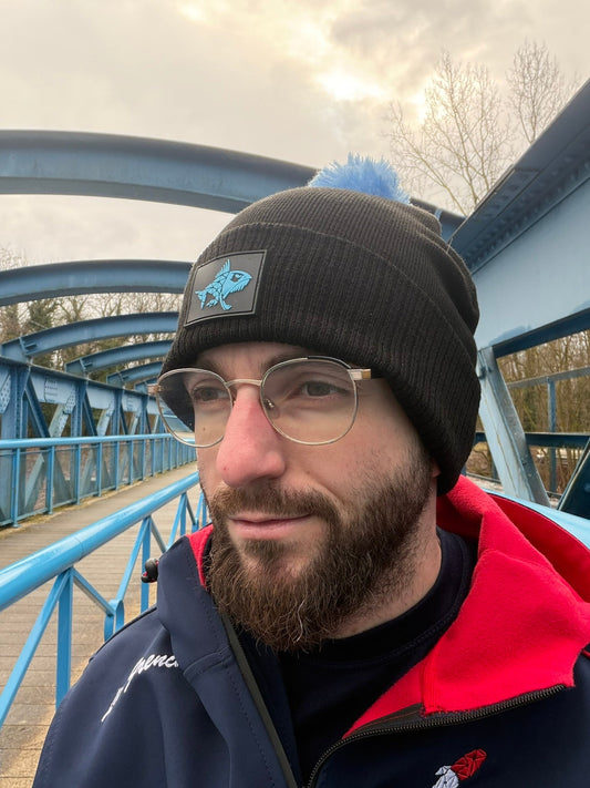 Homme portant un bonnet noir Connected Carper avec pompon bleu et logo poisson sur un pont bleu