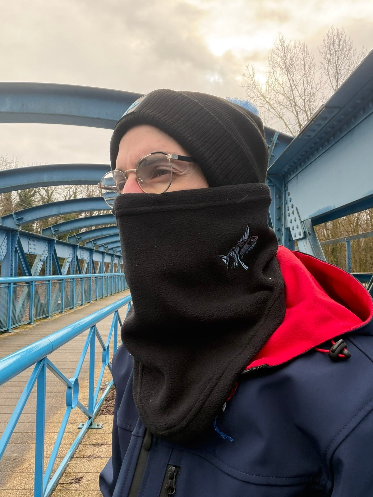 Homme portant un tour de cou polaire noir Connected Carper avec broderie de poisson sur un pont bleu en extérieur
