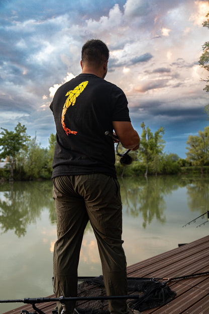 Connected Carper black Heritage t-shirt with orange carp logo on back, worn by a fisherman on a wooden pier at sunset.