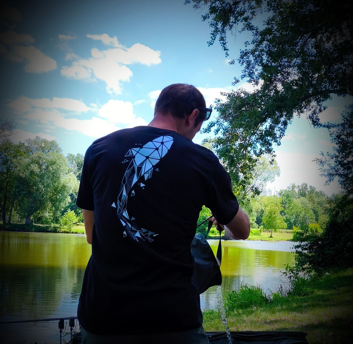 Connected Carper black t-shirt with a large white geometric carp graphic on the back, worn by a man at a lake.