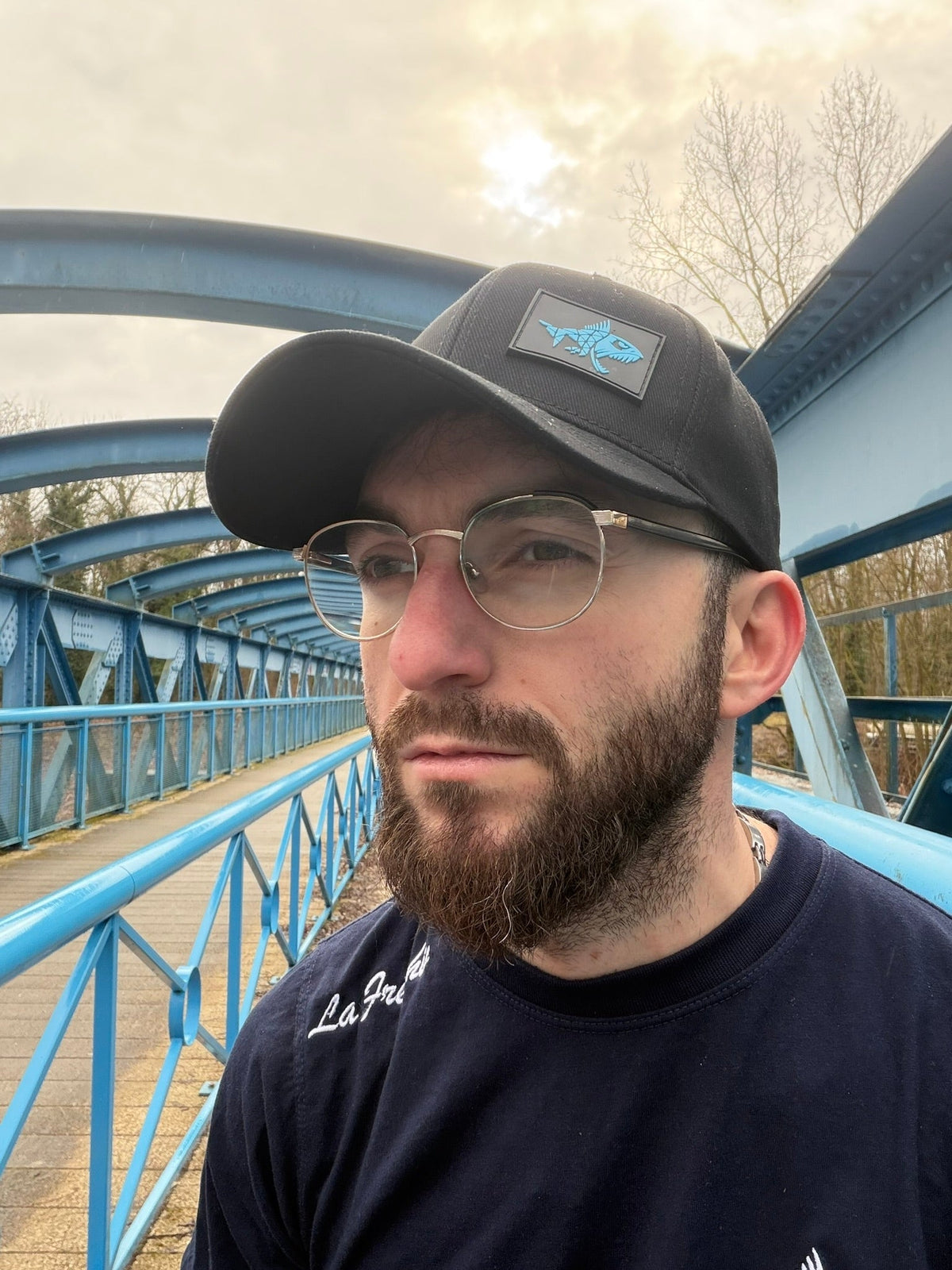Homme portant une casquette Connected Carper Predator noire en coton avec un patch en silicone bleu sur un pont