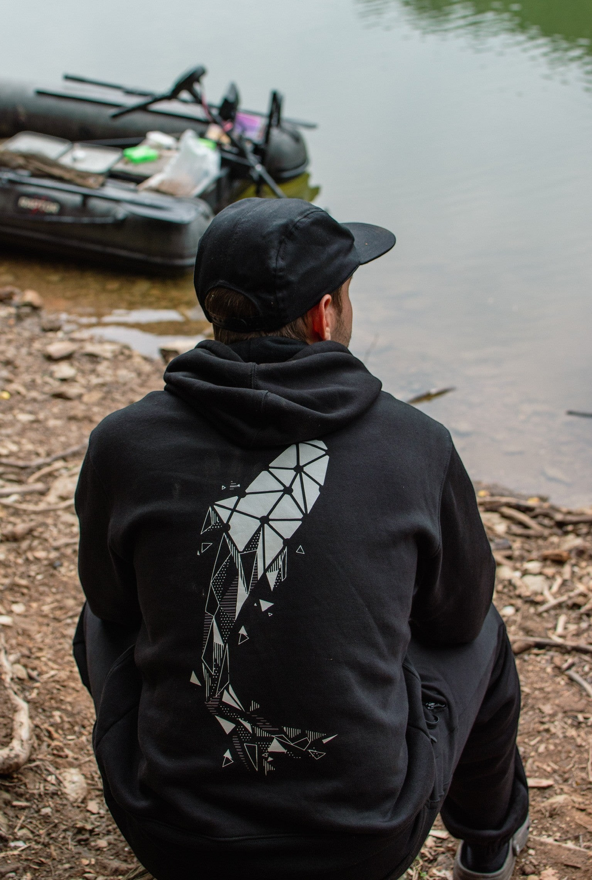 Connected Carper black hoodie with geometric carp print on back, worn by a fisherman by the water.