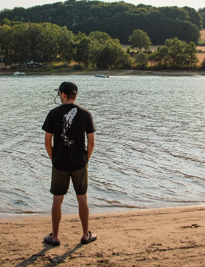 Connected Carper black t-shirt with geometric carp print on back, worn by a man standing by a lake at sunset.