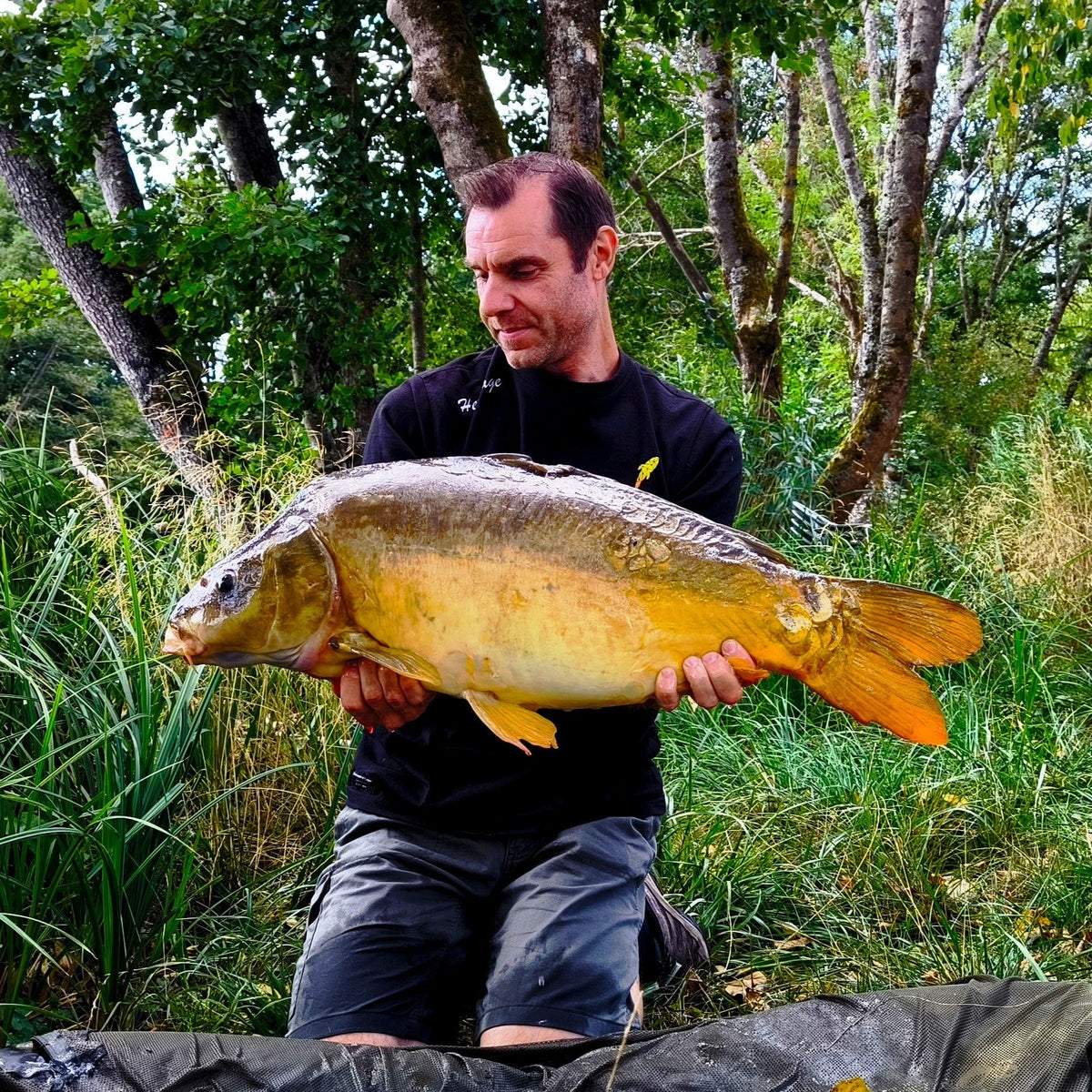 Connected Carper Heritage black t-shirt worn by a man holding a large carp fish by a lake.