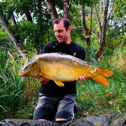 Connected Carper Heritage black t-shirt worn by a man holding a large carp fish by a lake.