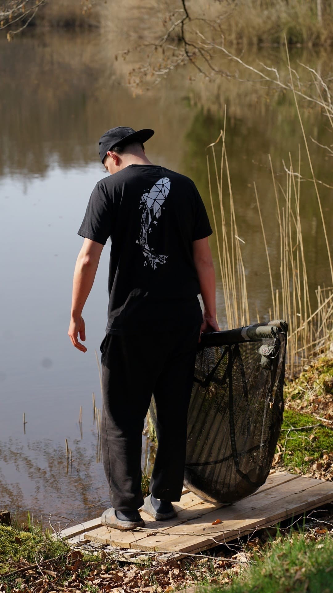 Homme portant un jogging noir Connected Carper avec cordon de serrage, debout au bord d'un étang avec une épuisette
