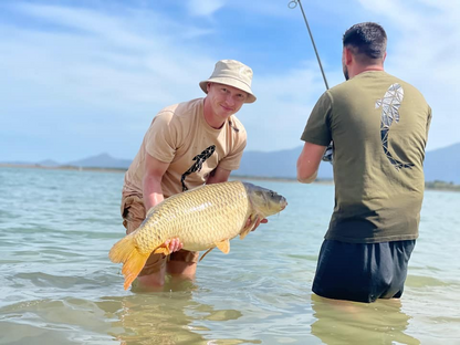 Connected Carper khaki fishing t-shirt worn by a man holding a large carp in a lake with another angler nearby