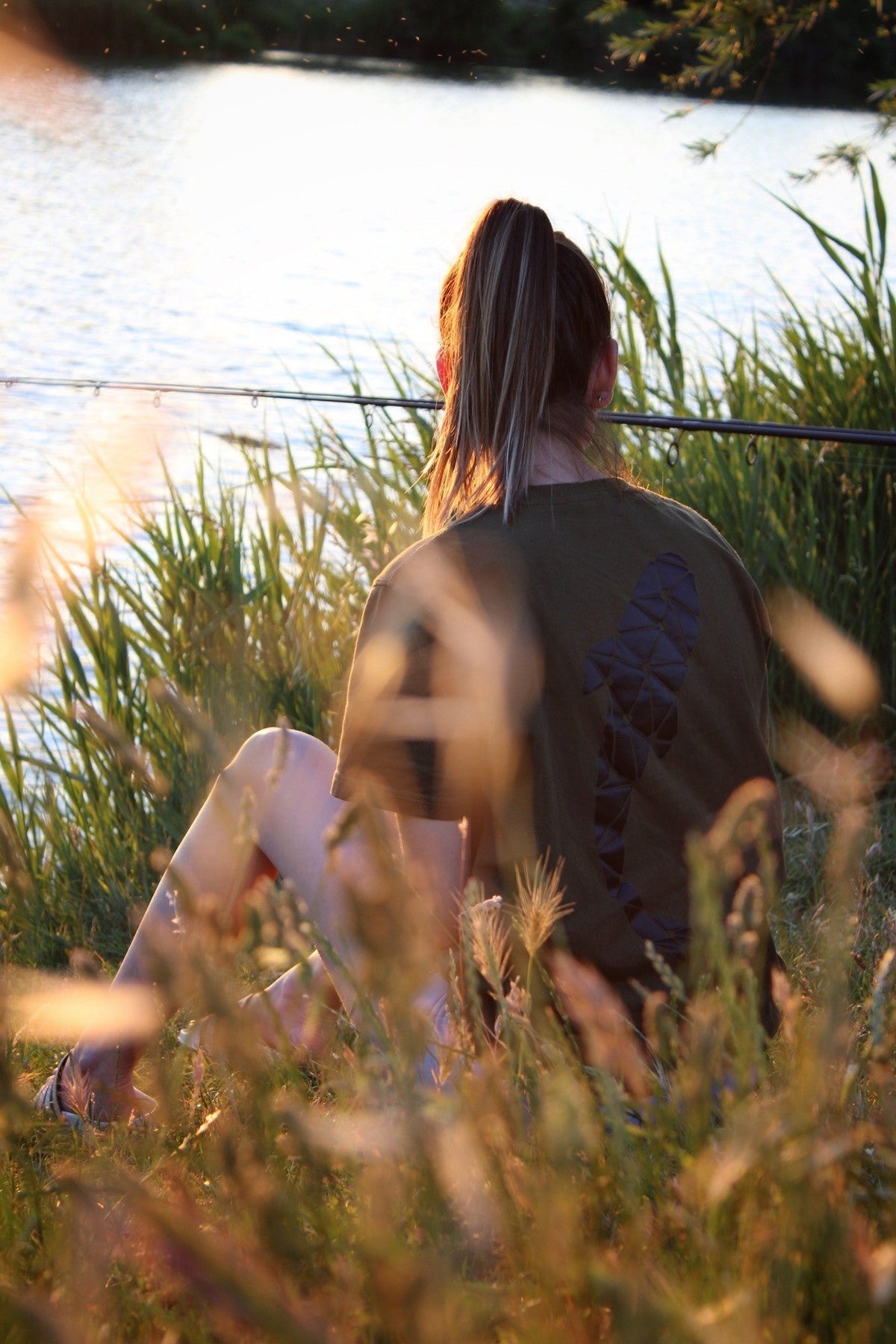 Connected Carper khaki fishing t-shirt with back logo worn by a woman sitting by a lake at sunset