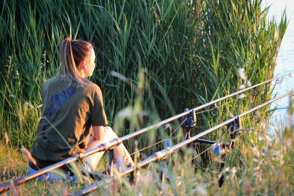 Connected Carper khaki fishing t-shirt worn by a woman sitting by a lake with fishing rods at sunset