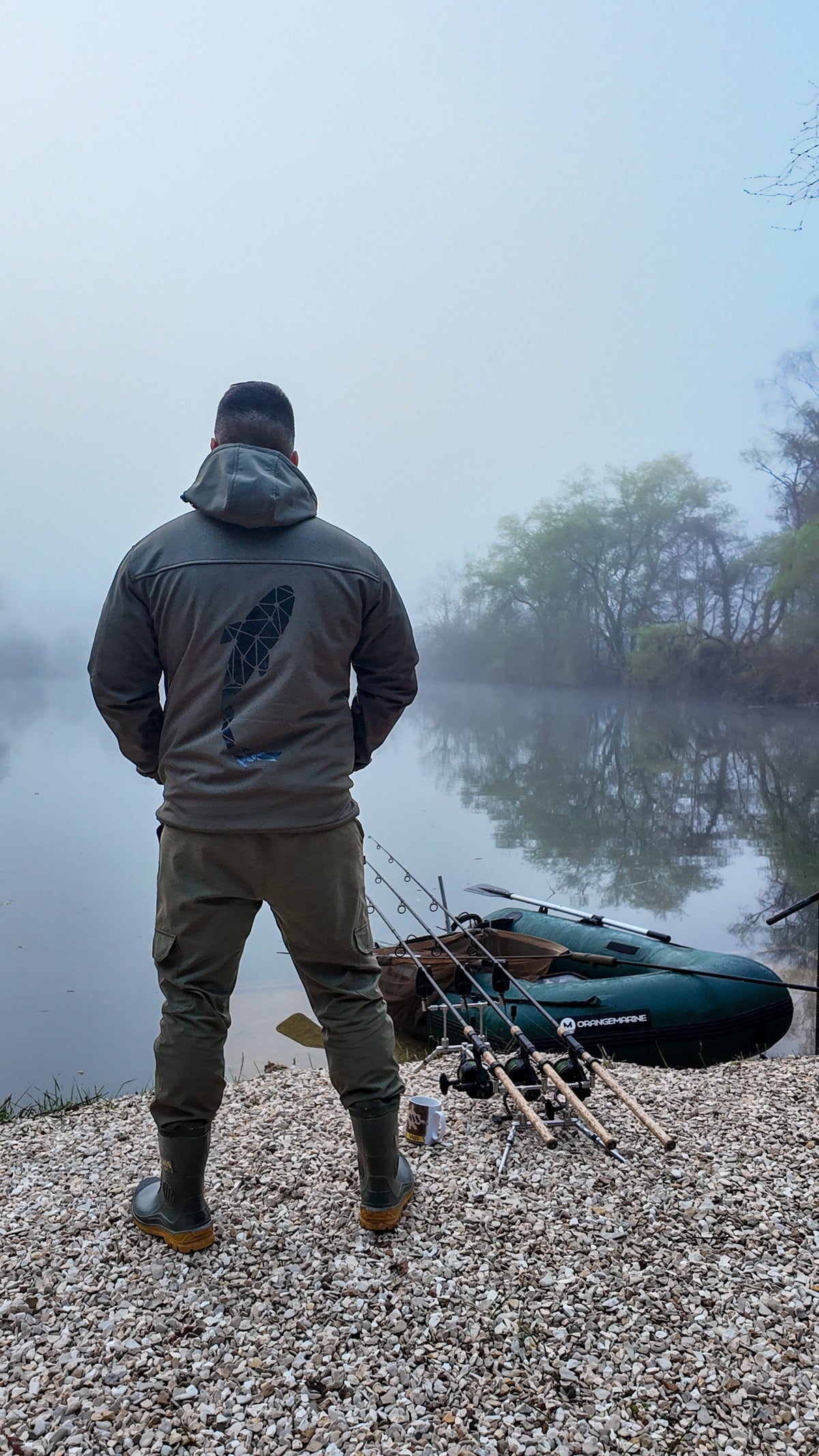 Connected Carper khaki softshell jacket with back logo, worn by a fisherman standing by a foggy lake with gear.