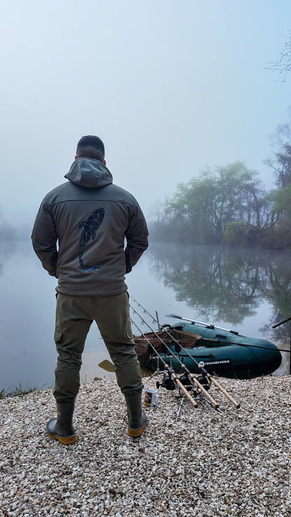Connected Carper khaki softshell jacket with back logo, worn by a fisherman standing by a foggy lake with gear.