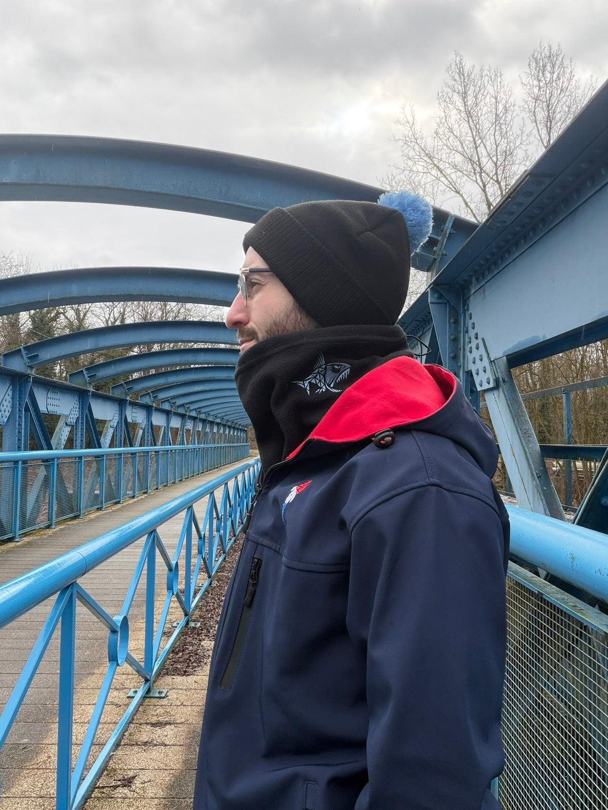 Homme portant un tour de cou polaire noir Connected Carper avec broderie de poisson sur un pont bleu en extérieur